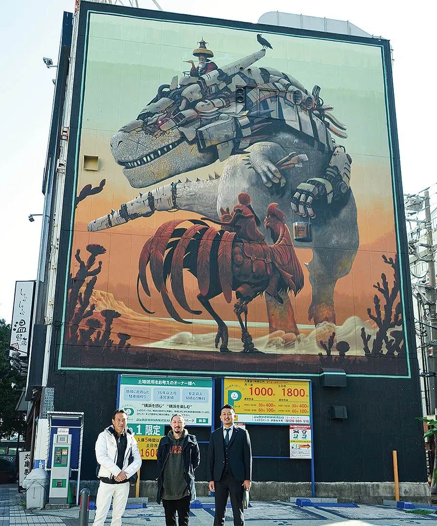 吉田町交差点近く、永興ビルの壁面に描かれた巨大壁画（写真は左から吉田理事長、Kensukeさん、山下さん）