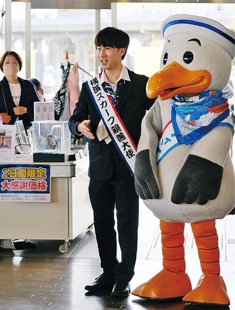 男性初のスカーフ親善大使となった渡辺悠希さん（19）と中区のマスコットキャラクター・スウィンギー。スカーフを身に付け登場（同実行委員会提供）